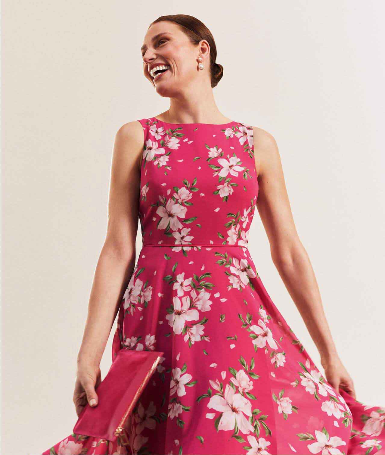 Hobbs model wears a pink floral midi dress with a matching clutch bag.