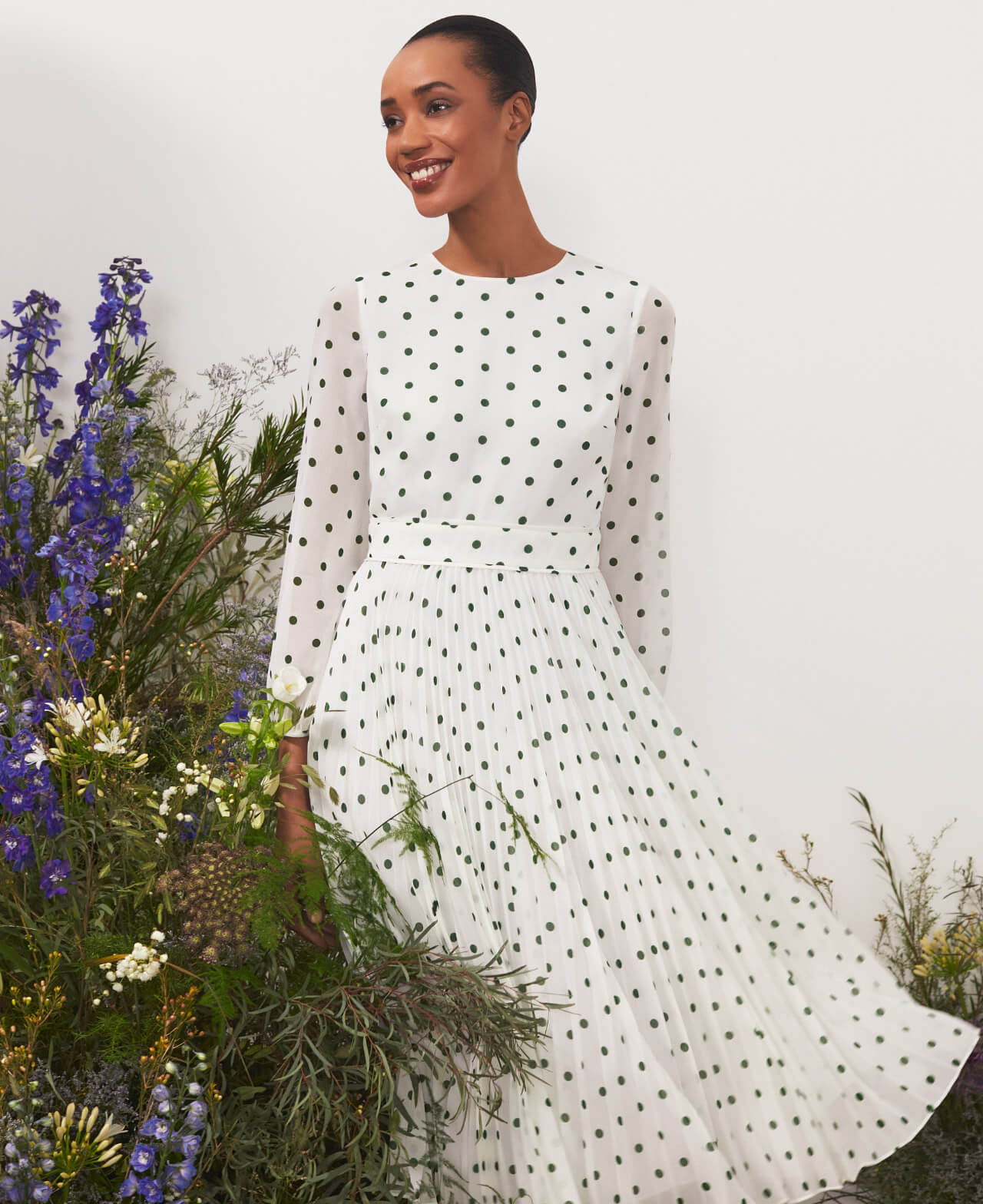 Hobbs stands next to a floral display wearing a polka dot fit and flare dress.