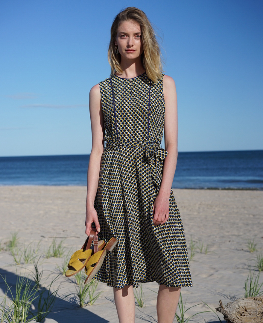 Iris poses in a blue geometric sleeveless dress with yellow suede sandals in hand on the beach.