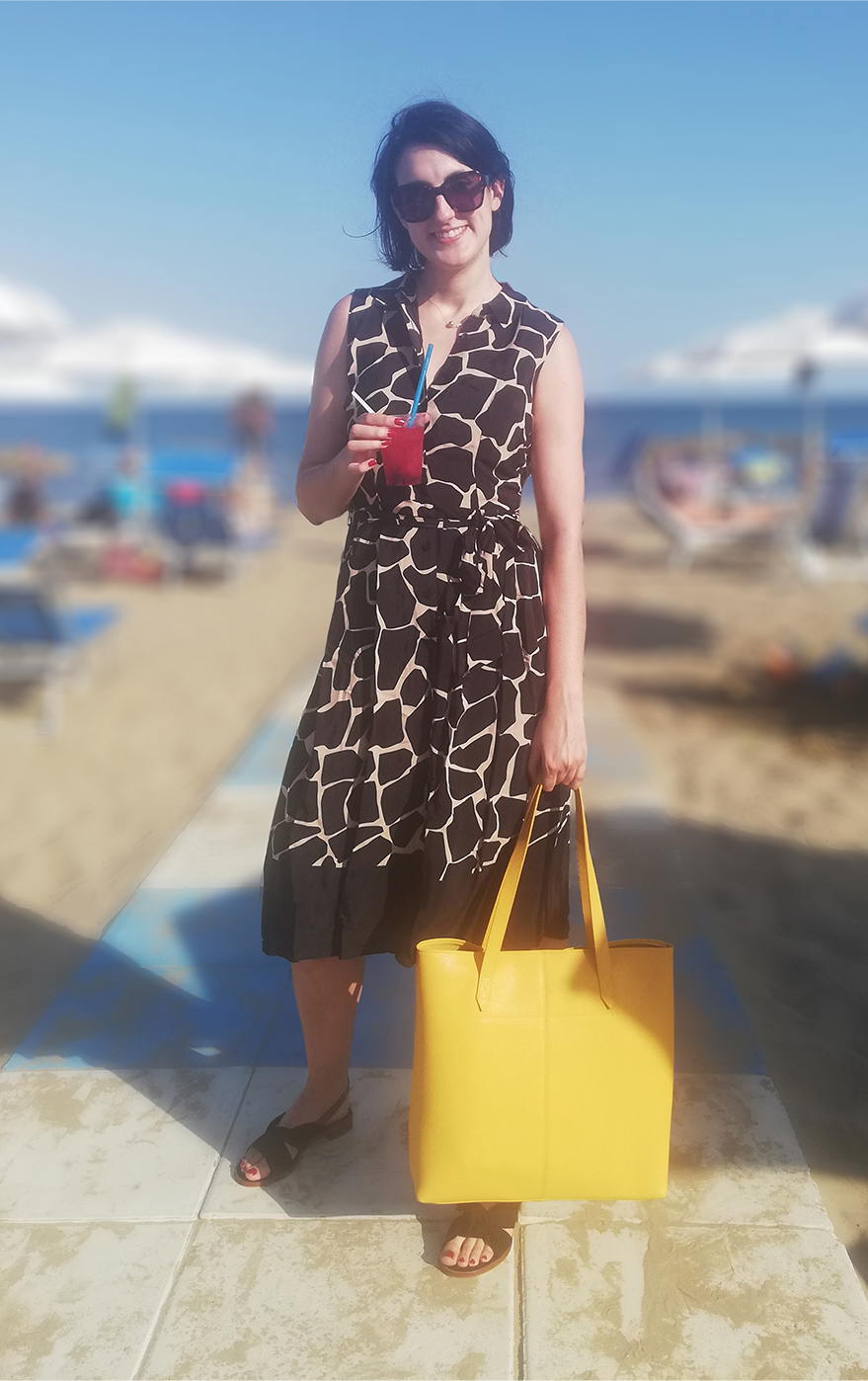 Marta Bonfaci, Senior Ecommerce Assistant, stands by the seaside holding a drink while wearing a sleeveless fit and flare shirt dress with an animal print, styled with a yellow tote bag and black slingback sandals.