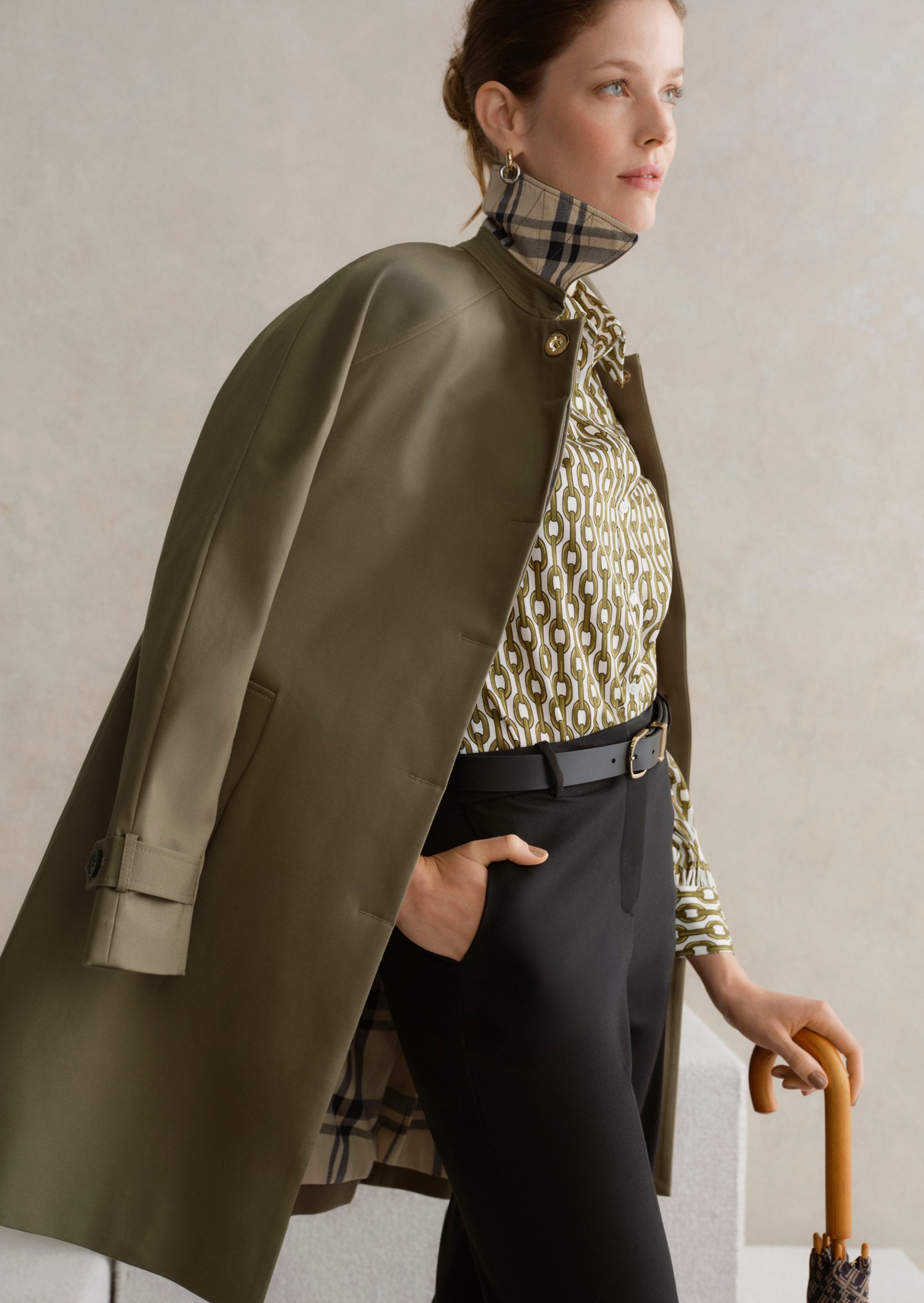 A gallery of two images displaying a model wearing a khaki trench coat, printed blouse and tailored trousers.