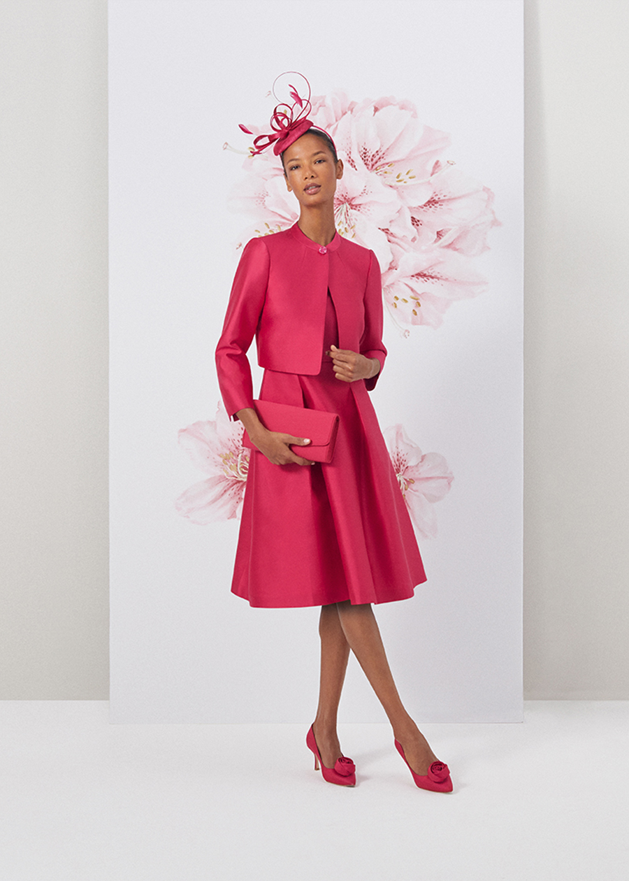 Image of model standing in front of a floral painted background wearing a matching fuschia pink jacket and dress two piece outfit.