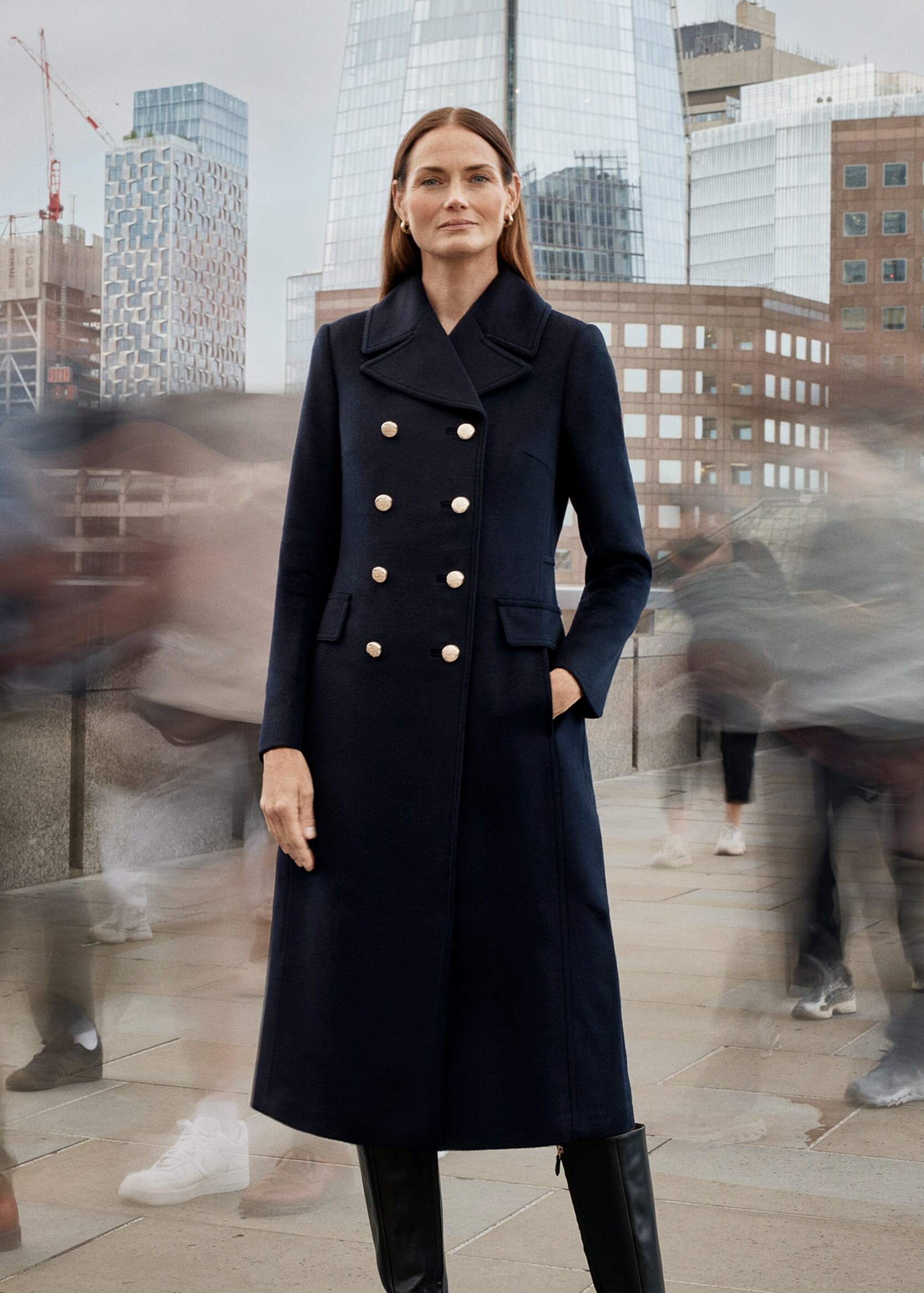 A model in a military navy coat stands still on a street as people rush past.