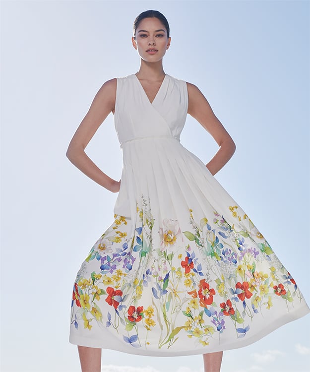 Woman poses in an elegant white midi dress with a floral motif along the bottom edge